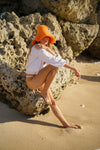 Model posing on a rock at the beach in Bloom Crochet Sun Hat, in Tangerine Orange 